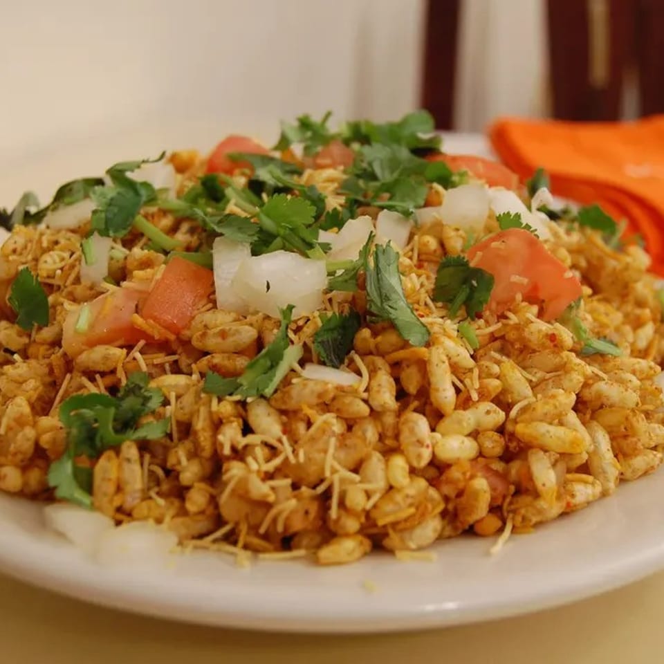 Plate of spicy puffed rice snack topped with sev, chopped tomatoes, onions, and fresh cilantro.