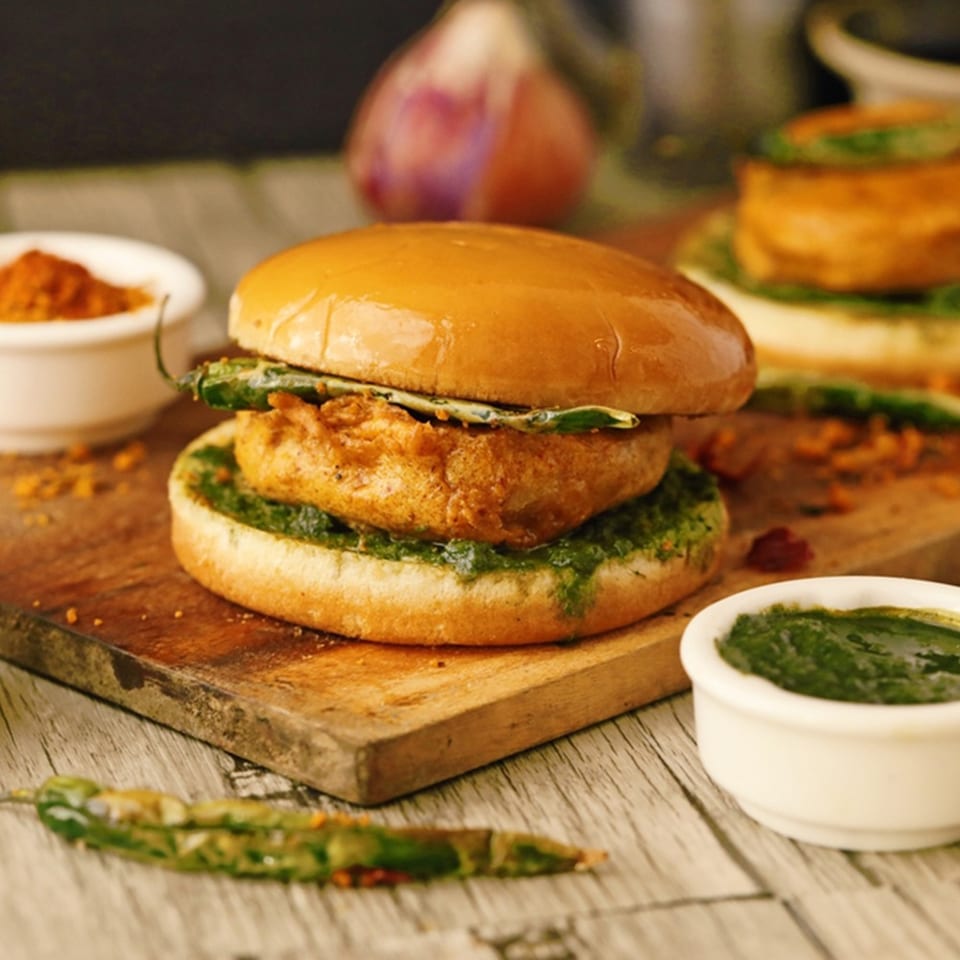Crispy chicken burger with green sauce and leafy greens on a wooden board, accompanied by dipping sauces and another burger.