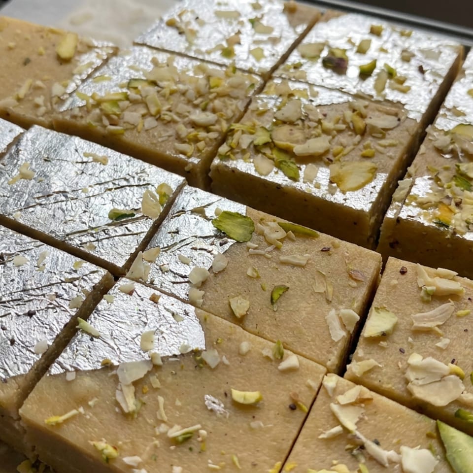 Square pieces of Indian milk fudge topped with edible silver leaf, sliced almonds, and pistachios on a tray.